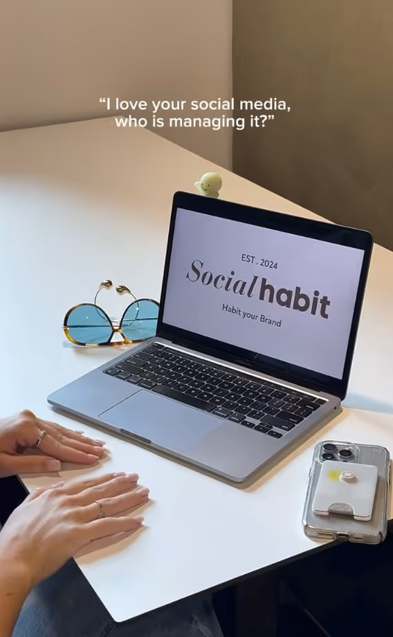 Laptop on a white desk displaying 'Social habit EST. 2024 Habit your Brand', with hands resting nearby, sunglasses, and a phone also on the desk.